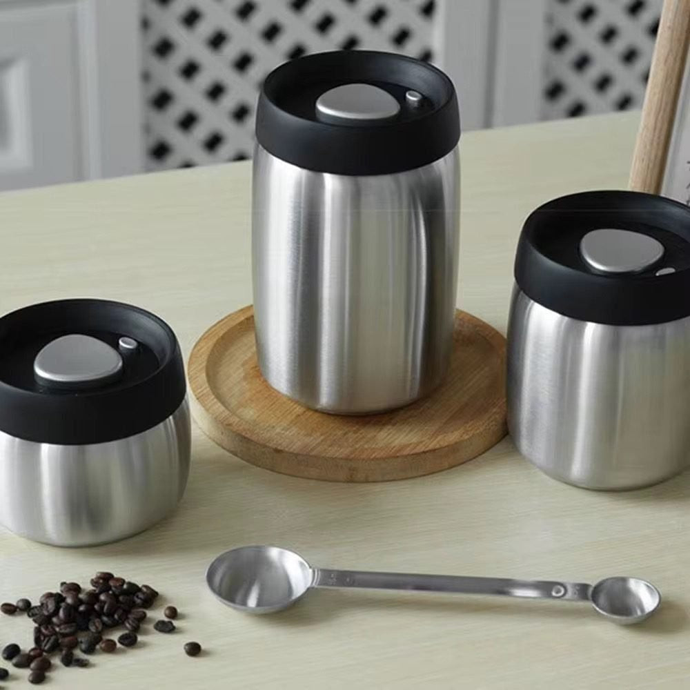 Stainless steel vacuum coffee canisters with pump lids and a scoop on a wooden board